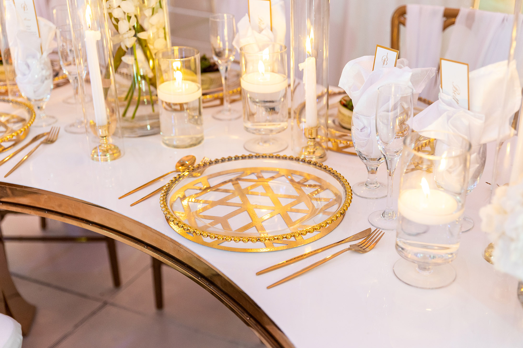White flower arrangements and floating candles on a table with gold accents on plate settings, silverware, tables and chairs eccessories by ellen www.eccessoriesbyellen.com