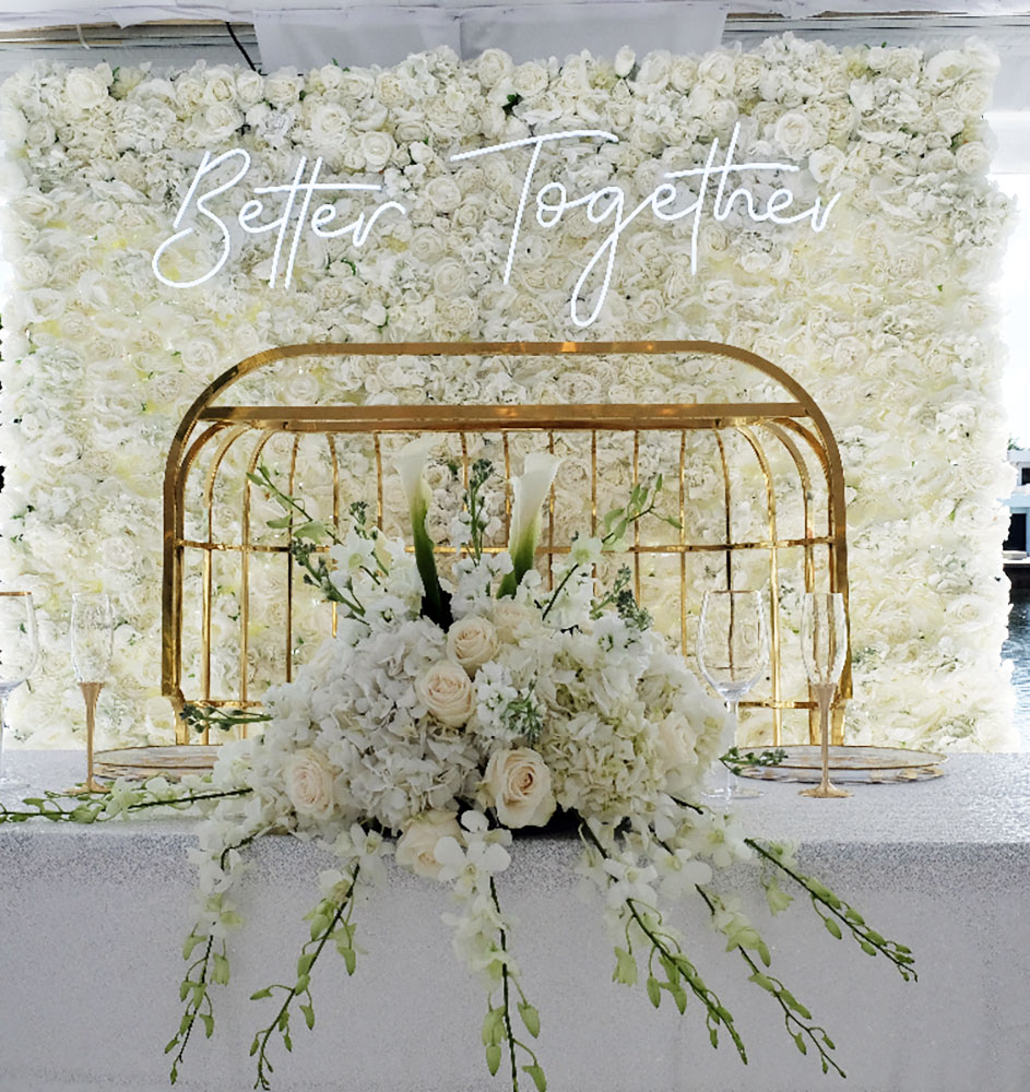 A wedding reception setup by eccessories by ellen. Display made up of center display of a white floral arrangement on table with gold accents in front of a Better Together white neon sign with flower backdrop display eccessories by ellen www.eccessoriesbyellen.com